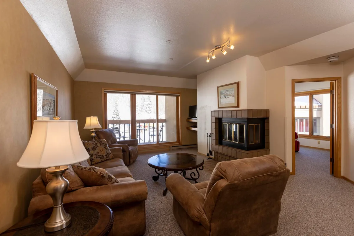 The image showcases a warm and inviting living space with a glowing fireplace, plush sofa set, and expansive windows revealing snowy mountain views—perfect for relaxing with friends or family after a day on the slopes.