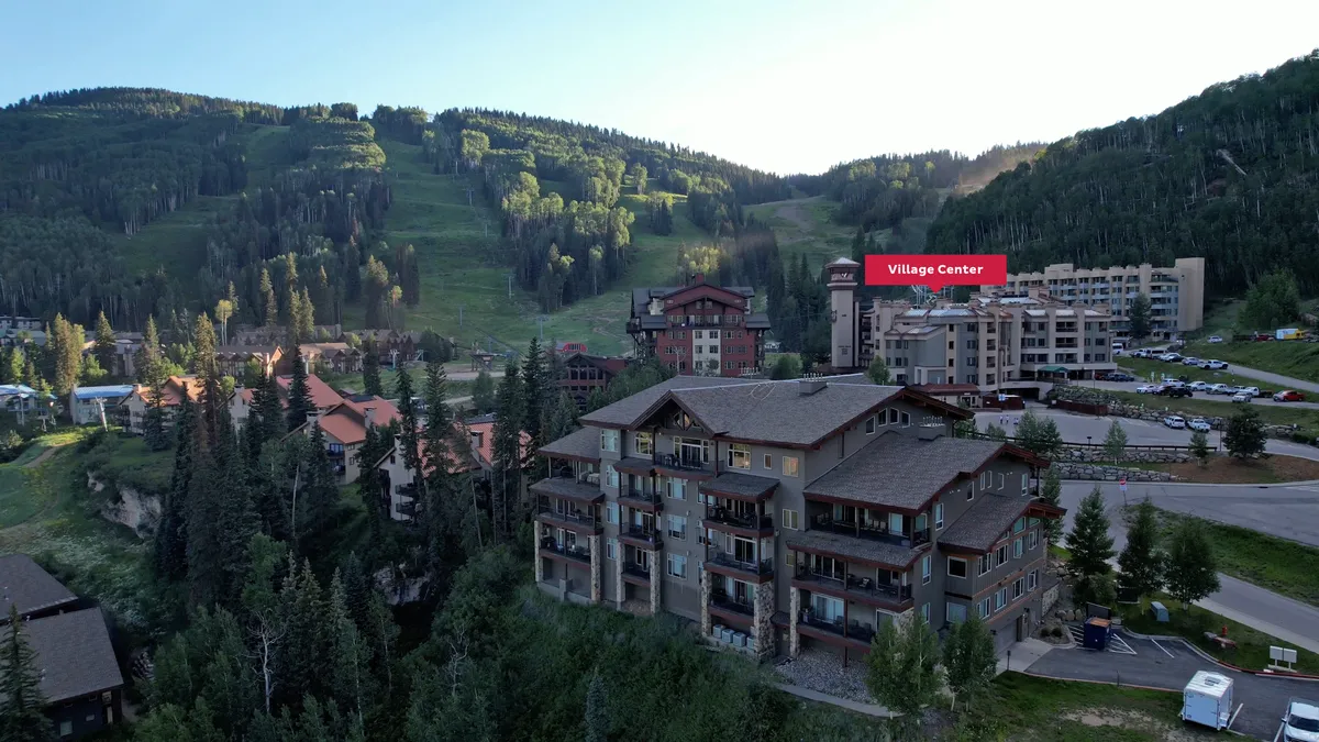 An exterior view of the charming mountain village, featuring wooden architecture nestled at the base of dramatic peaks. The village center bustles with potential activities, while lush trees frame the scene, hinting at both adventure and serenity.
