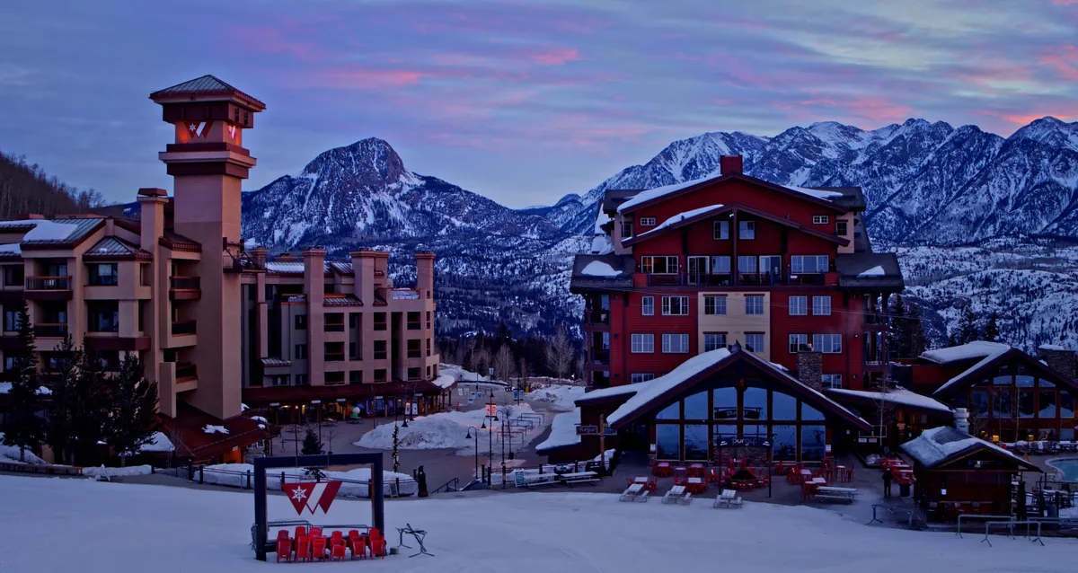 A wintery exterior scene featuring the Purgatory Resort buildings nestled amid snow-covered ground, framed by majestic mountains in the background. Skiers and snowboarders are visible, highlighting the ski-in/ski-out experience. The scene is set at sunset, casting a golden glow over the landscape and inviting a sense of adventure and serenity.