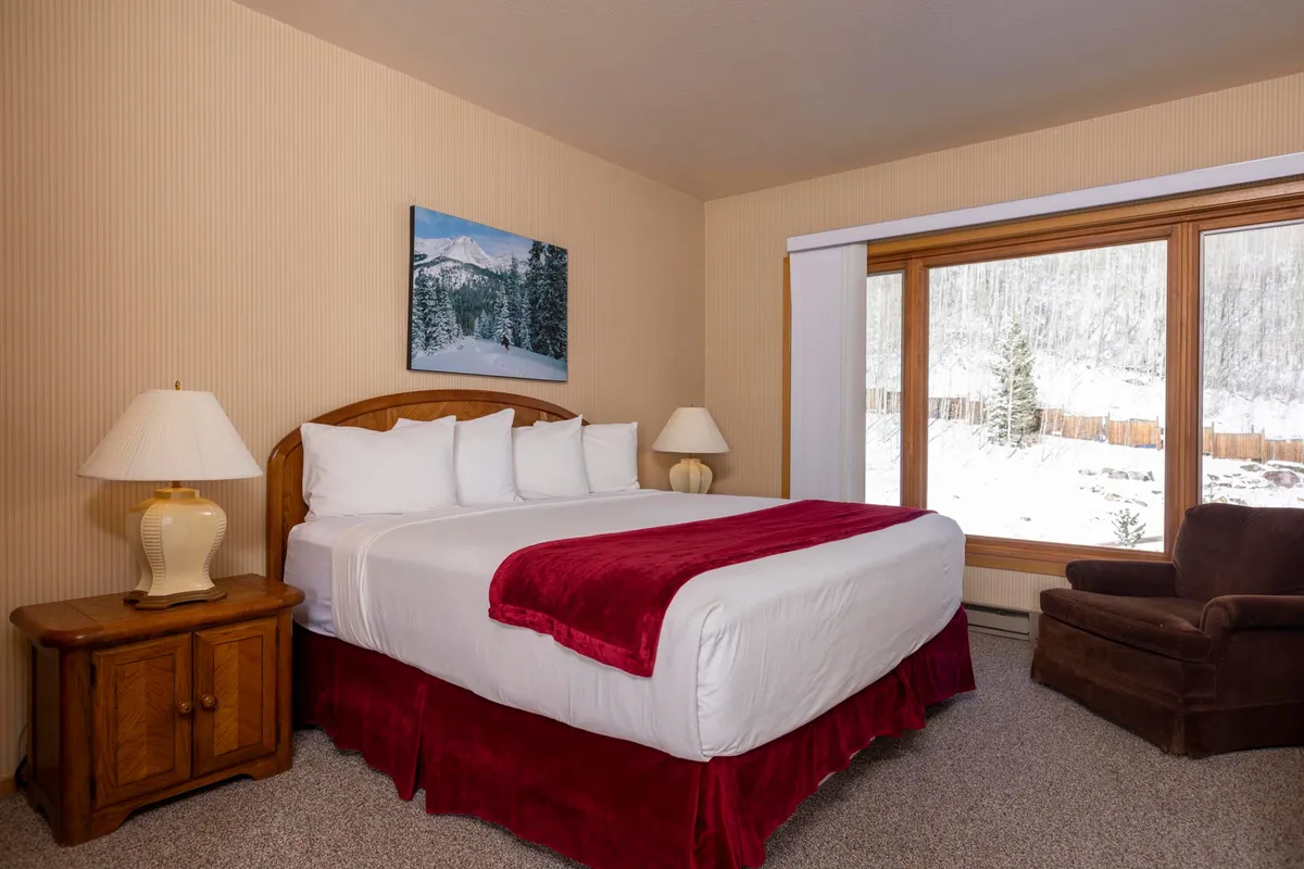 A warm, inviting bedroom with a large bed draped in a red blanket, snowy mountain artwork, a window view of a winter wonderland, a wooden nightstand with lamp, and a cozy armchair by the window.
