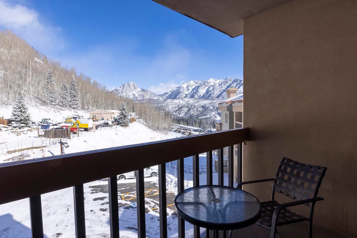 A serene balcony seating area overlooks a breathtaking mountain landscape, with snow-covered trees stretching out toward majestic peaks. The space is inviting, perfect for enjoying a morning coffee or quietly contemplating the natural beauty.