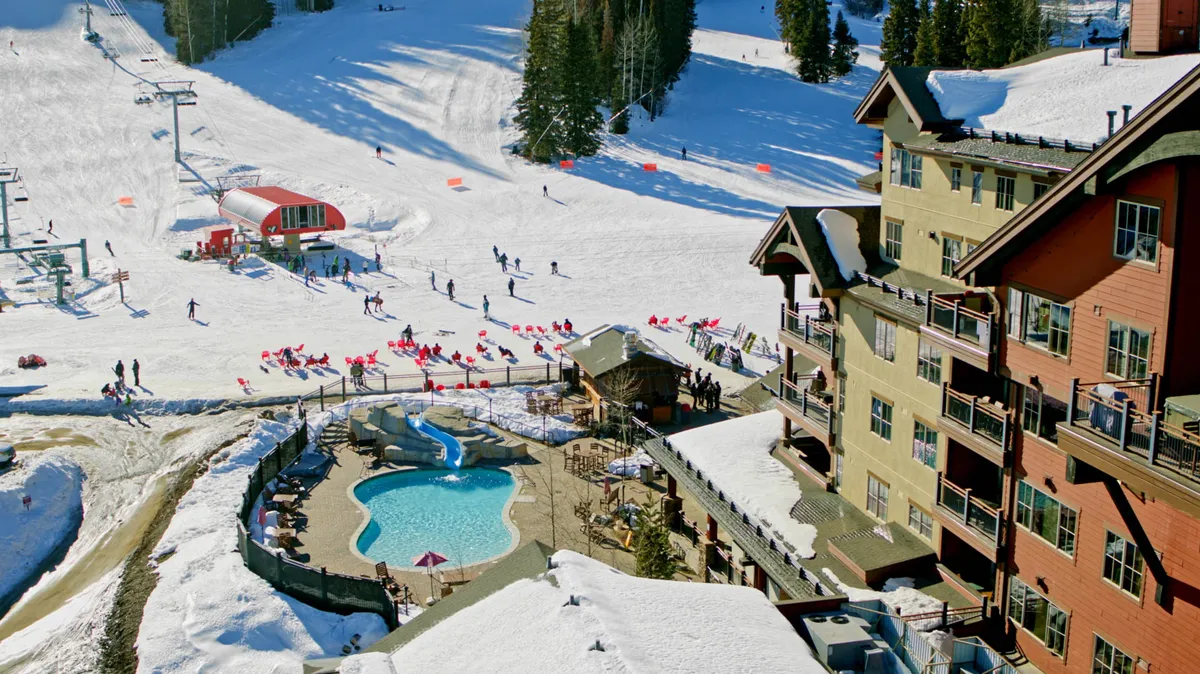 A lively exterior shot featuring ski slopes, a swimming pool, a ski lift, and a lodge. People are skiing and socializing near the pool and hot tub, set against the backdrop of snow-capped mountains and a glowing lodge at sunset.