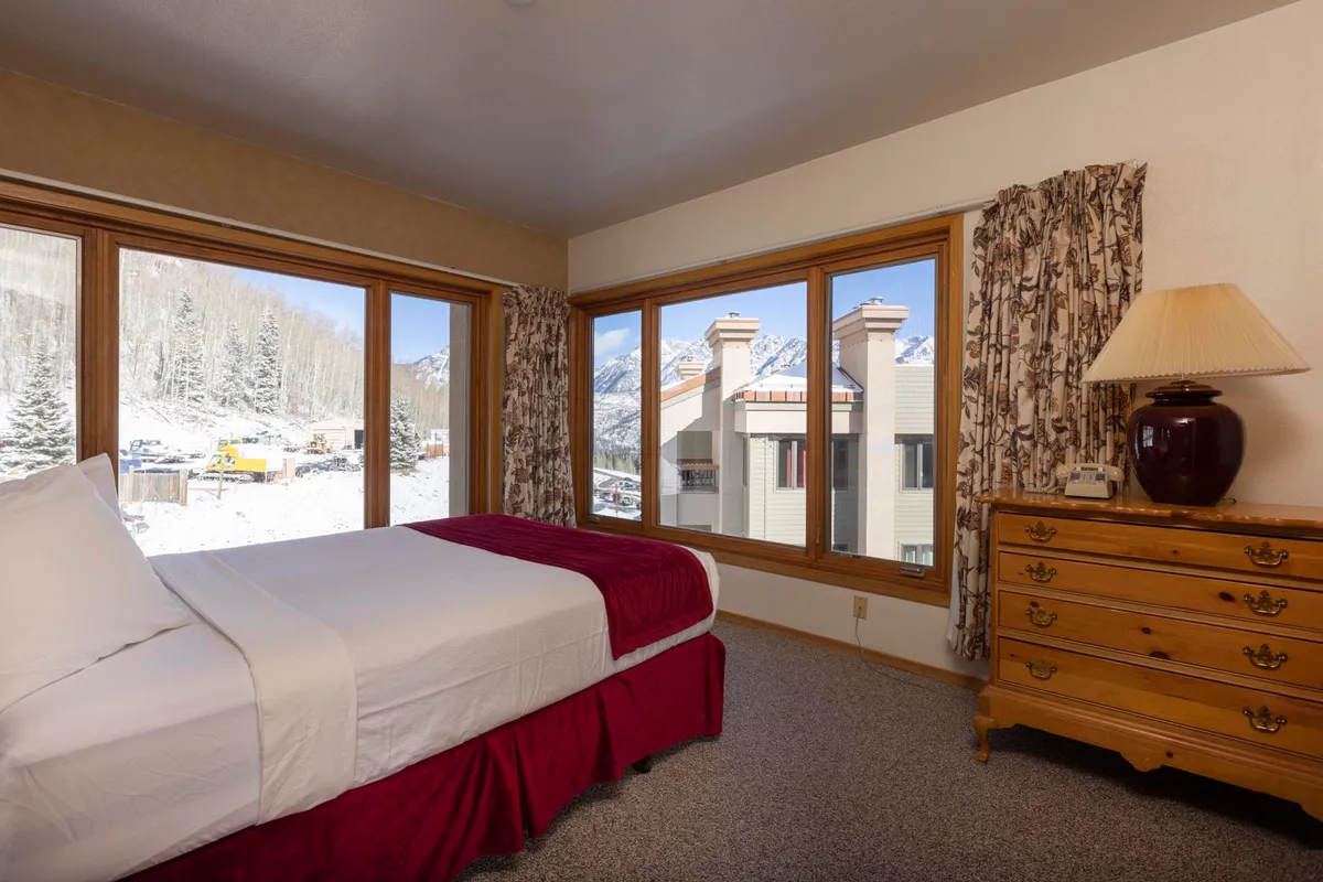 A serene master bedroom featuring a plush bed with crisp white linens and a burgundy accent, large windows showcasing stunning mountain views, and a wooden dresser with a lamp. Soft lighting and a tranquil color palette create a restful atmosphere.