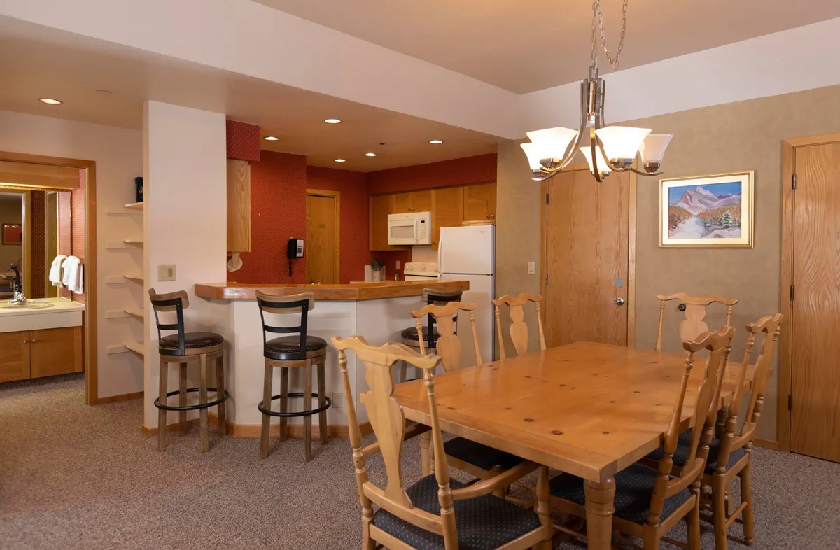 A stylish dining area with a large table set beneath an elegant chandelier, adjacent to a modern kitchen counter. The space is adorned with tasteful artwork and designed to accommodate family dinners or group gatherings.