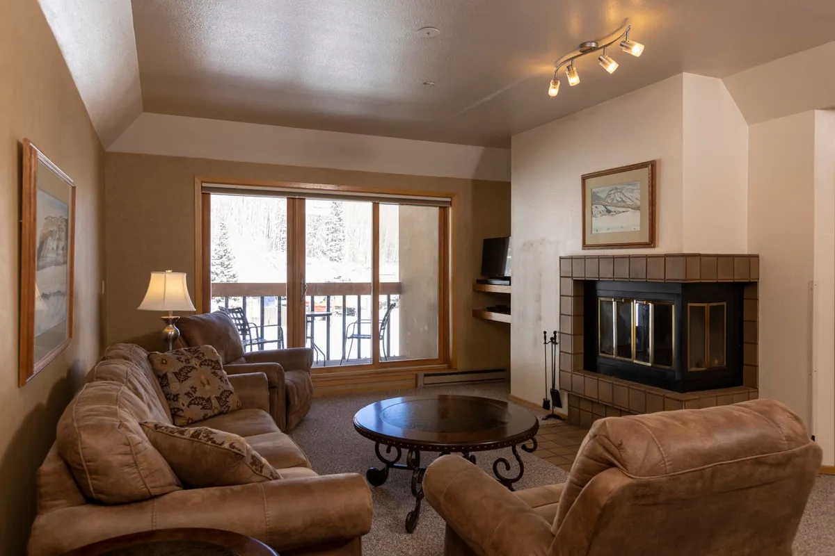 A warmly-lit living area featuring a comfortable sofa set positioned around a fireplace, with large windows that provide sweeping views of the snowy landscape outside. The space is designed for socializing and relaxation, with a coffee table at the center and ample natural light pouring in.