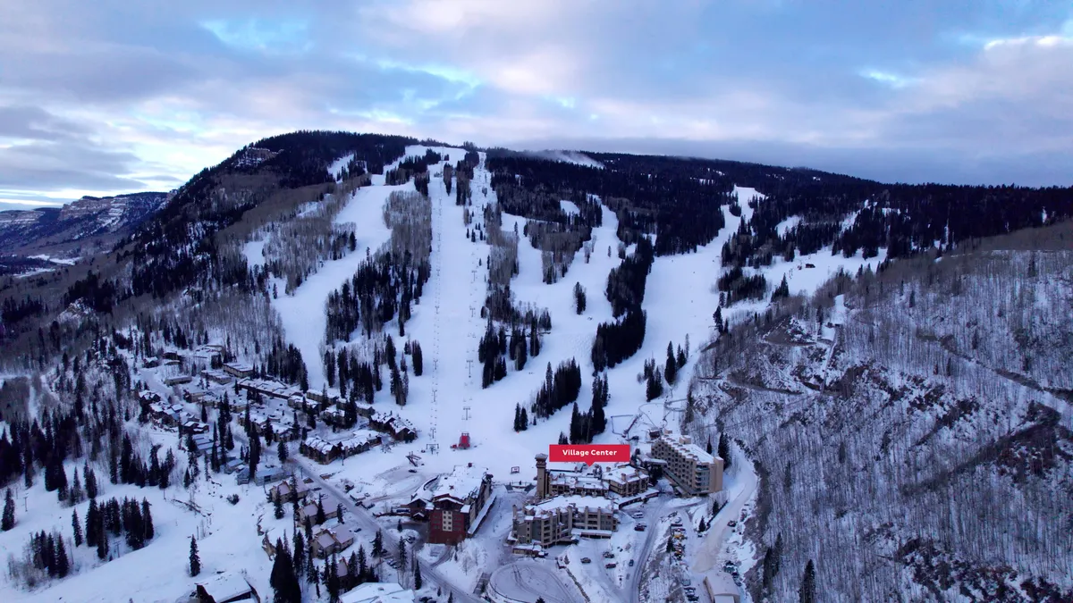 A panoramic exterior view showcasing ski slopes directly adjacent to the vibrant Village Center, surrounded by a lush forest. The scene is alive with skiers and snowboarders enjoying the mountain, with the sun setting over the peaks and the village bustling below.
