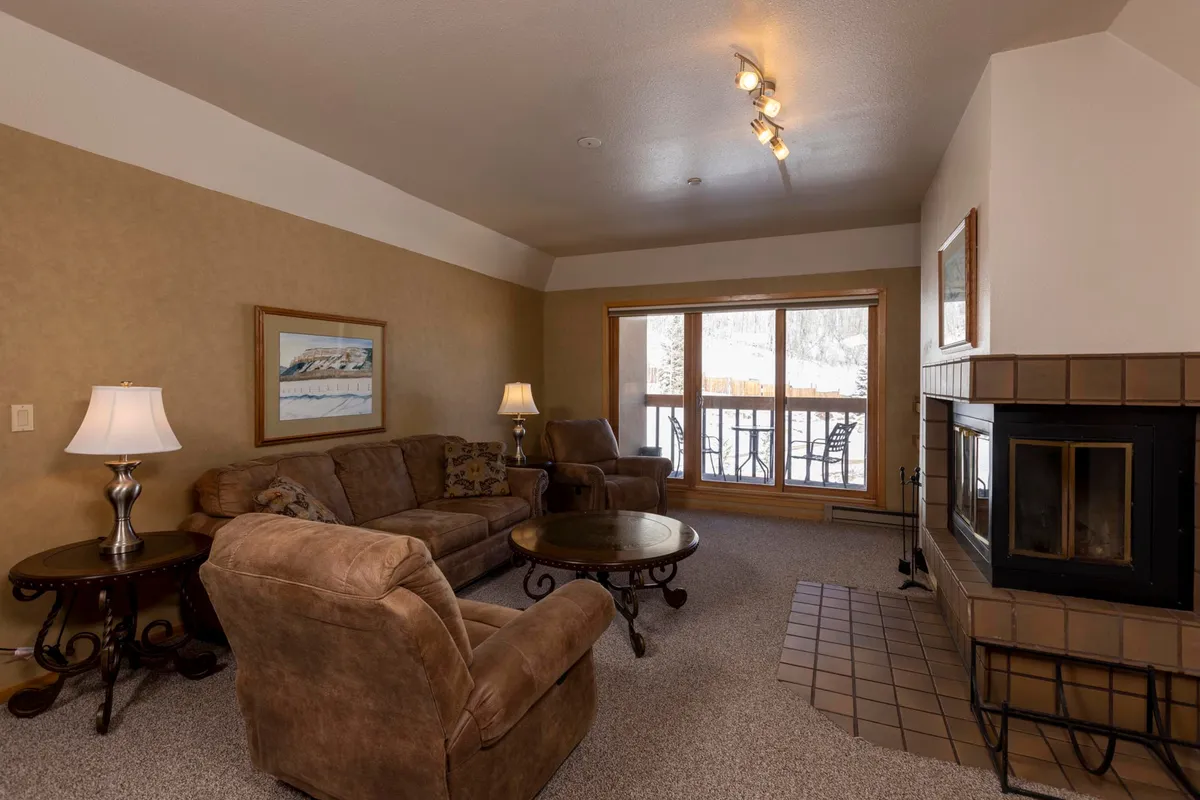 A close-up view of the living room’s plush sofa, coffee table, and a glowing fireplace. A large window frames a snowy outdoor scene, enhancing the indoor-outdoor connection. Soft lighting creates an inviting and intimate ambiance.