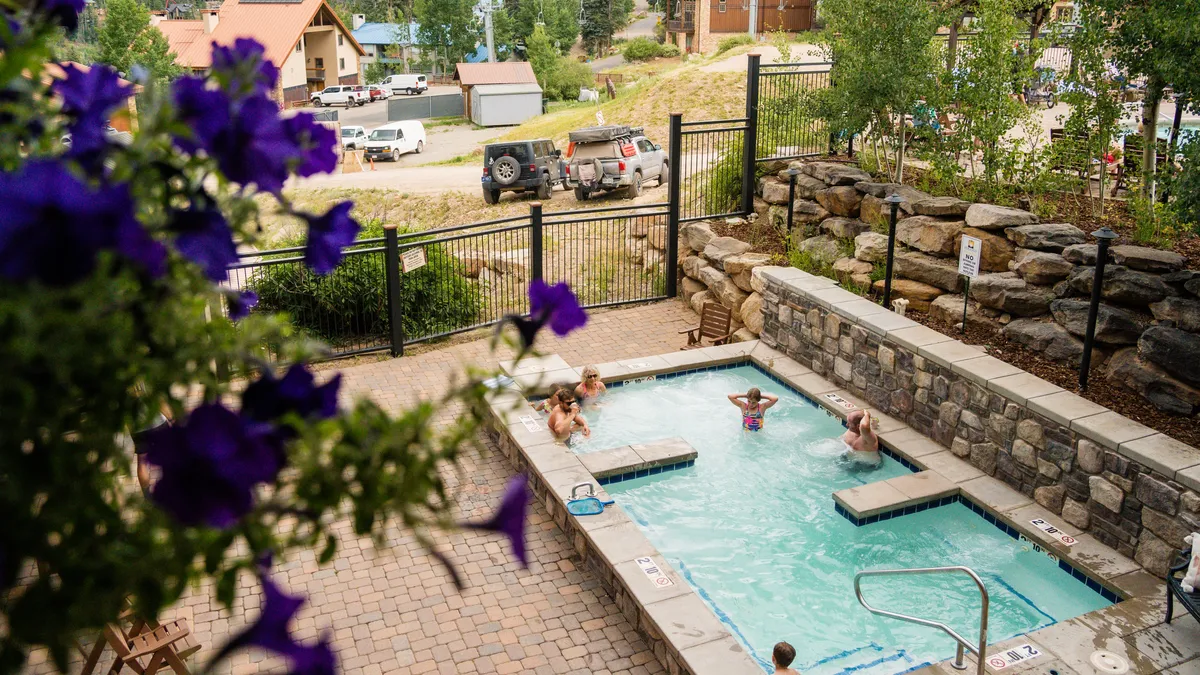 A sparkling outdoor swimming pool bordered by colorful flowers, with people enjoying the water and lounging poolside. The surrounding area feels vibrant and social, suggesting a cheerful and leisurely atmosphere perfect for both families and couples.