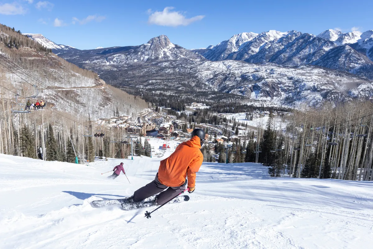 Skiers in vibrant jackets zip down snowy slopes with the ski resort village, chairlift, and snow-capped peaks in clear view. The energetic action, expansive scenery, and visible amenities underscore the thrill and convenience of slope-side adventure.