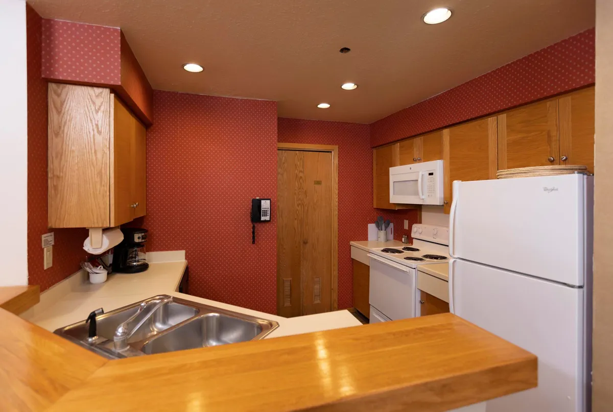 An inviting kitchen scene focusing on a kitchen sink, stove, refrigerator, and wooden countertops, with homey wallpaper in the background. The layout encourages gathering, meal preparation, and socializing.