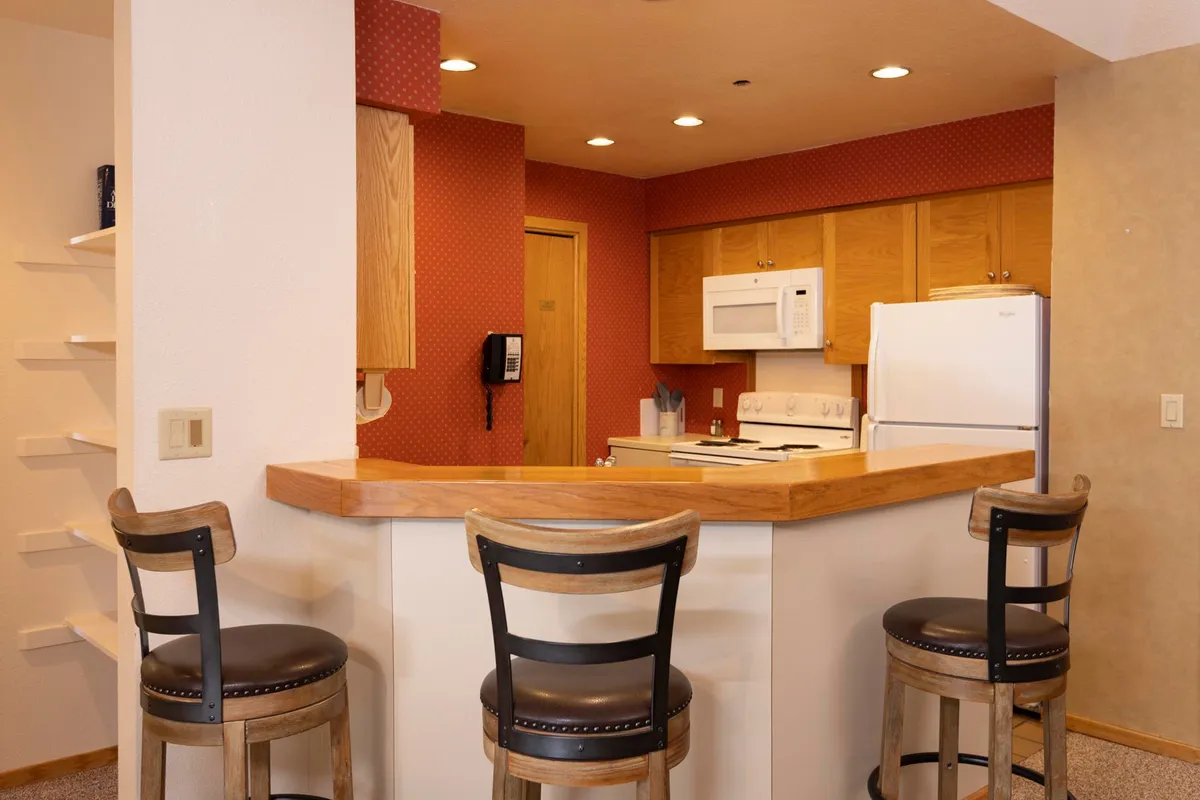 A welcoming kitchen interior featuring a bar counter with stools, modern kitchen appliances, wooden cabinetry, and stylish light fixtures. The space has a warm and inviting atmosphere, ideal for both cooking and entertaining.