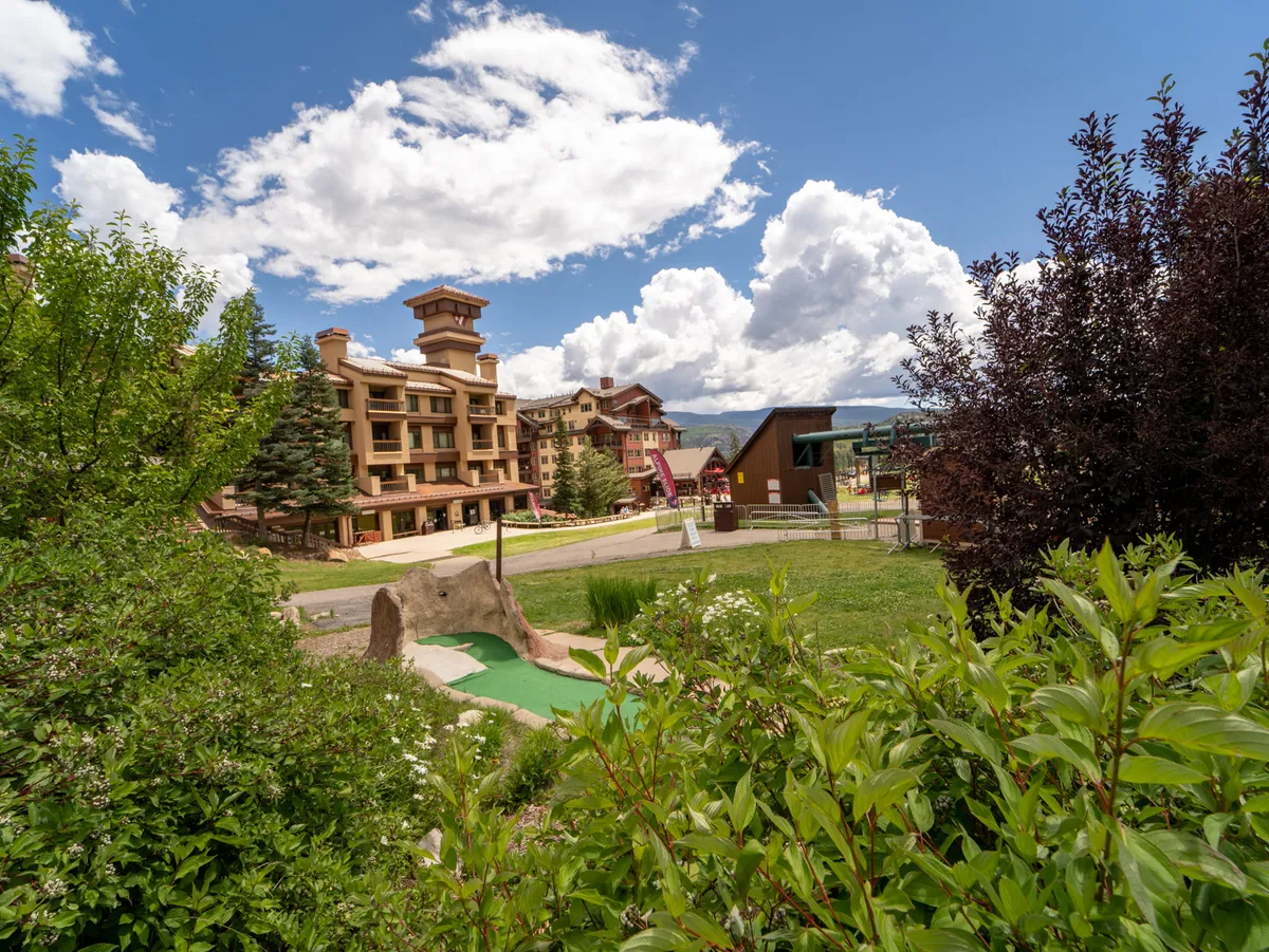 A cheerful exterior scene featuring the resort’s multi-story condo building, a mini-golf course, and lush greenery. The space is filled with families and friends enjoying outdoor games and dining, set against a backdrop of blue skies and mountains.